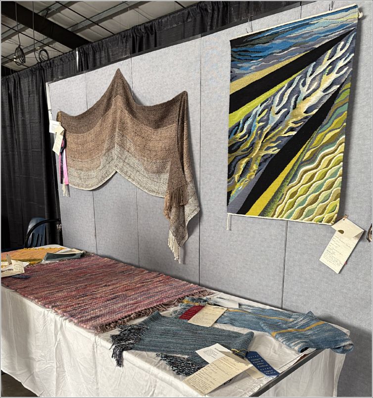  The Fiber Arts Gallery included this lovely weaving display. The top right piece is a beautifully woven tapestry; cotton warp and wool weft. Theres a brown tone shawl on the wall to its left, a rag rug on the table next to two finely woven scarves. Photo: Linda Bolewicz.